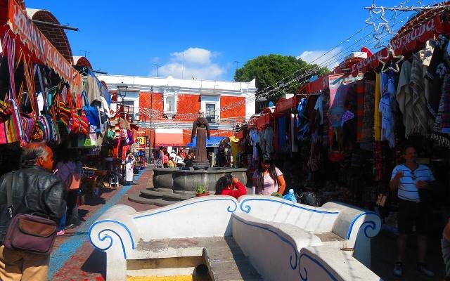 Craft stalls hung with embroidered clothes around a tiled fountain at El Parián market